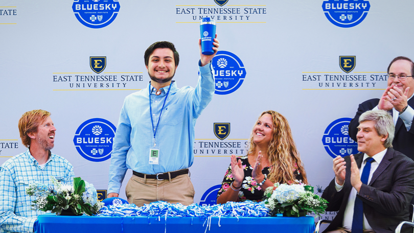 A BlueSky student being awarded and lifting a branded travel coffee mug