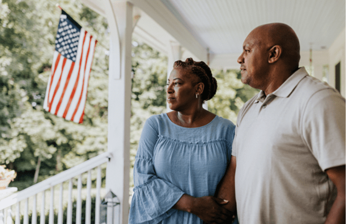 Senior couple standing on their porch with an American Flag in the background.